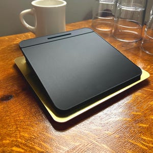 May include: A black rectangular tray with a textured surface sits atop a gold-colored tray. A white mug and several clear drinking glasses are in the background. The items are on a wooden table.