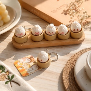 May include: A collection of dumpling-shaped keychains. Each keychain features a white dumpling with a smiling face, nestled in a small, tan-colored bamboo steamer. The keychains are arranged on a wooden tray, with one keychain and a tag in the foreground.