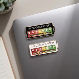 May include: Two enamel pins in the shape of a battery, each with the text "MY SOCIAL BATTERY". The pins feature a slider with a range of emoji faces from sad to happy, and a lightning bolt. One pin has a black border, the other white.