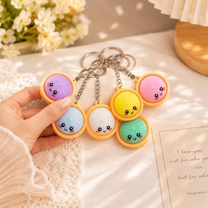 May include: A collection of colorful keychains featuring round, smiling faces in various colors: purple, blue, white, yellow, pink, and green. Each keychain has a silver chain and a yellow border. The keychains are a fun and cheerful accessory.