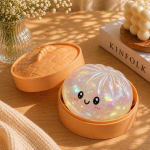 May include: A pearlescent, dumpling-shaped stress ball with a smiling face, nestled in an orange steamer basket. The lid of the basket is beside it. The background includes a vase of flowers and a book.