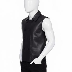 May include: A black leather vest with a double-breasted design. The vest has a notched collar and four visible buttons. The mannequin is wearing black jeans. The vest appears to be sleeveless.