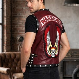 May include: A maroon leather vest featuring a black trim with white diamond-shaped studs. The back displays a large embroidered patch with the word WARRIORS in red letters above a graphic of a skull with wings and flames in red, white, and yellow.