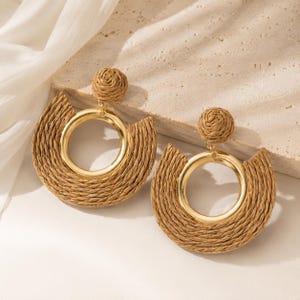 Könnte beinhalten: Ein Paar Ohrhänger mit einem runden, geflochtenen beigen Strohstecker, der mit einem goldfarbenen Metallring verbunden ist. Der Ring ist teilweise von einem halbmondförmigen, geflochtenen Strohrahmen umschlossen, was für eine Textur sorgt.