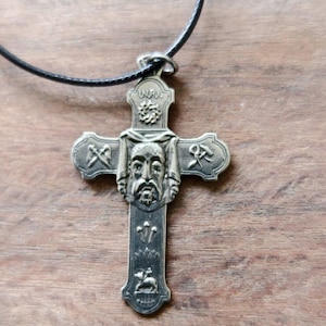 Puede incluir: Collar con colgante plateado con un diseño de cruz detallado. La cruz tiene una cara central, símbolos y la inscripción "INRI" en la parte superior. Cuelga de un cordón negro.