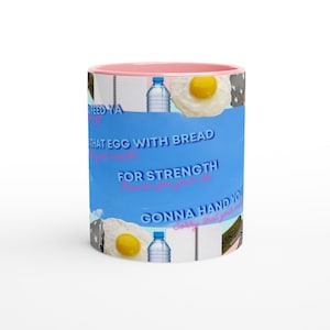 May include: A ceramic mug with a pink interior and rim, featuring a collage design on a blue background. The print includes images of fried eggs, water bottles, and spiral notebooks, with text reading "FEED YA", "THAT EGG WITH BREAD", "FOR STRENGTH", and "GONNA HAND YOU".