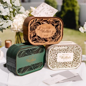 May include: Three oval wooden card boxes with laser-cut leaf patterns. The top box is natural wood with 'Theodore & Claire Walker May 20, 2026'. The green box features 'Walker' in gold, and the white box displays 'Marco & Emma Walker May 20, 2026'.