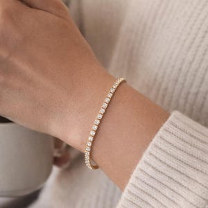 May include: A gold tennis bracelet featuring a line of clear, sparkling gemstones. The bracelet is displayed on a wrist, with a soft, neutral background. This bracelet is a timeless piece of jewellery.
