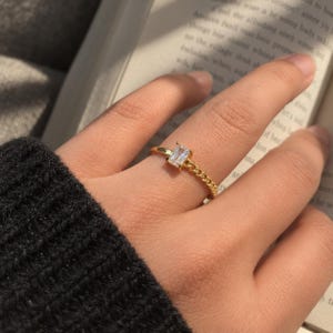 Puede incluir: Un anillo de tono dorado que presenta una piedra transparente única de corte baguette rectangular montada en cuatro garras, conectada a una delicada banda de cadena metálica que rodea el dedo, mostrando un acabado pulido y superficies reflectantes.