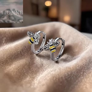 Peut inclure: Une paire de boucles d'oreilles créoles argentées avec un motif d'abeille. Les abeilles ont des rayures noires et jaunes et sont ornées d'une petite pierre transparente. Les boucles sont posées sur une surface beige.