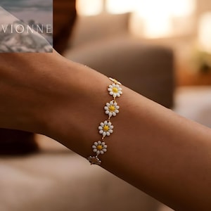 Könnte beinhalten: Ein goldfarbenes Armband mit Gänseblümchen-Anhängern mit weißen Blütenblättern und gelben Zentren. Das Armband wird am Handgelenk getragen und zeigt das zarte Blumendesign. Das Armband ist ein Schmuckstück.
