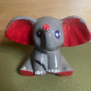 Può includere: Una piccola statuetta di elefante in ceramica lucida dipinta di grigio con grandi orecchie rosso brillante e piedi rossi. L'elefante ha occhi delineati in blu, una proboscide con punta rossa e un'illustrazione di una piuma nera sul petto.