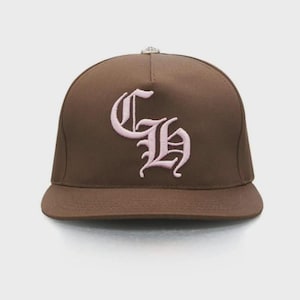 Puede incluir: Gorra de béisbol marrón con visera plana. La gorra presenta un diseño bordado en rosa en la parte delantera, posiblemente iniciales o un monograma. La gorra es de color liso y tiene un ajuste estructurado.