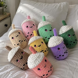 Puede incluir: Una colección de peluches de té boba hechos a mano con ganchillo en varios colores, incluyendo rosa, amarillo, verde y marrón. Cada peluche presenta una tapa de ganchillo blanca, una pajita pequeña y rasgos faciales bordados. Los peluches están colocados sobre una colcha blanca.