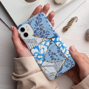 Op de afbeelding: Een blauwe en witte telefoonhoes met een geometrisch patroon van bloemen en gouden accenten. De hoes heeft een camera-uitsparing en een witte achtergrond.