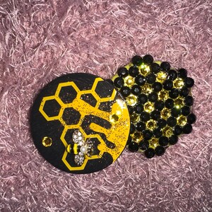 May include: Two decorative round coasters. One black coaster features a yellow honeycomb design with a jeweled bee. The other coaster is covered in black and gold gems. The coasters are on a pink, fuzzy surface.