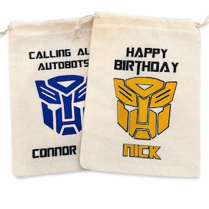 Könnte beinhalten: Zwei beigefarbene Kordelzugbeutel mit schwarzem Text und farbenfrohen Grafiken. Auf einem Beutel steht "CALLING ALL AUTOBOTS" mit einem blauen Logo und dem Namen "CONNOR". Auf dem anderen steht "HAPPY BIRTHDAY" mit einem gelben Logo und dem Namen "NICK".