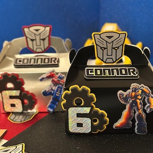 May include: Two gift boxes with Transformer-themed decorations. One box is silver with a red accent, and the other is black with a yellow accent. Each box has a name and the number 6. Additional Transformer character cutouts are visible.