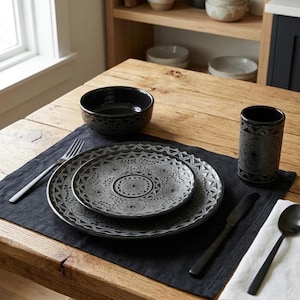 May include: A dark gray ceramic dinnerware set featuring a detailed embossed floral and geometric pattern. The set includes a large dinner plate, a smaller salad plate, a deep bowl, and a tall tumbler, all arranged on a black linen placemat.