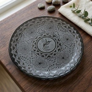 May include: A round, translucent gray glass plate featuring an embossed design. The center displays a hummingbird motif surrounded by a circular geometric border. The outer rim is decorated with repeating floral patterns and zigzag lines.