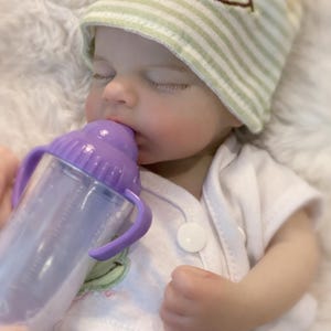 Op de afbeelding: Een babypop met gesloten ogen drinkt uit een paarse babyfles met handvatten. De baby draagt een witte onesie en een gestreepte groen-witte muts. De baby heeft een lichte huid met roze wangen.