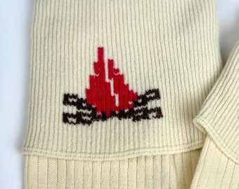 May include: Cream-colored knitted leg warmers with a red and brown embroidered design. The design appears to be a stylized flame or floral motif. The leg warmers are folded over at the top.