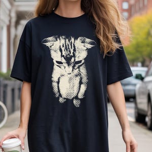 A high-contrast, vintage halftone graphic design on a dark shirt featuring the viral 'sad cat' meme image. The print depicts a small, cream-colored tabby kitten looking downward with a melancholy expression, flattened ears, and a sad face. The artwork has a distinct retro screentone texture with visible dot patterns, giving it a stylized, grunge-inspired look