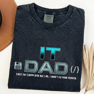 May include: Dark grey t-shirt with the text "IT DAD" in a blue and grey design, including a floppy disk icon and the phrase "SINCE THE FLOPPY DISK AGE | NO, I WON'T FIX YOUR PRINTER."