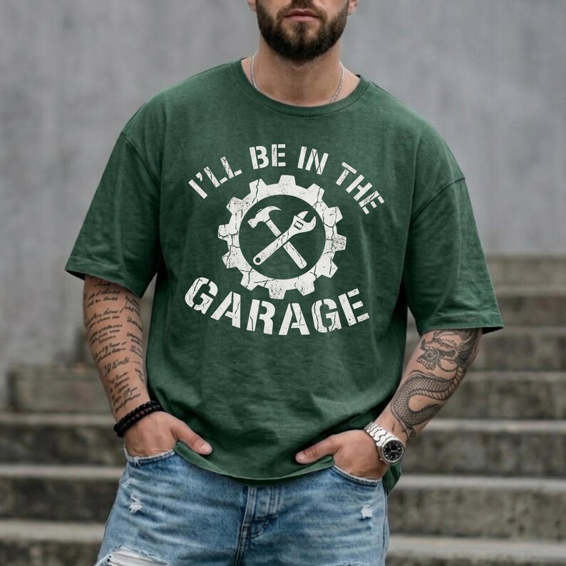 May include: Green t-shirt with the text "I'LL BE IN THE GARAGE" in white. The design features a gear with a hammer and wrench inside. The shirt is a solid green color.