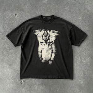 Peut inclure: Un t-shirt noir &agrave; manches courtes avec une impression graphique contrast&eacute;e centr&eacute;e repr&eacute;sentant un chaton. Le chaton est repr&eacute;sent&eacute; dans une couleur cr&egrave;me claire avec un motif textur&eacute; de style matriciel, montrant la t&ecirc;te et le haut du corps.