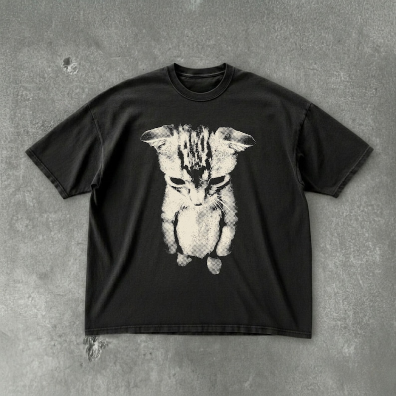 Peut inclure: Un t-shirt noir &agrave; manches courtes avec une impression graphique contrast&eacute;e centr&eacute;e repr&eacute;sentant un chaton. Le chaton est repr&eacute;sent&eacute; dans une couleur cr&egrave;me claire avec un motif textur&eacute; de style matriciel, montrant la t&ecirc;te et le haut du corps.