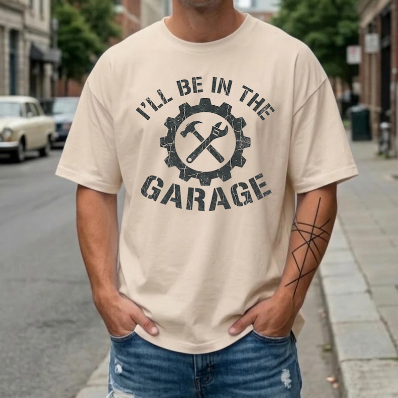 May include: Beige t-shirt with the text "I'LL BE IN THE GARAGE" in distressed black lettering. A gear graphic with a hammer and wrench is centered above the text. The shirt is a casual style.