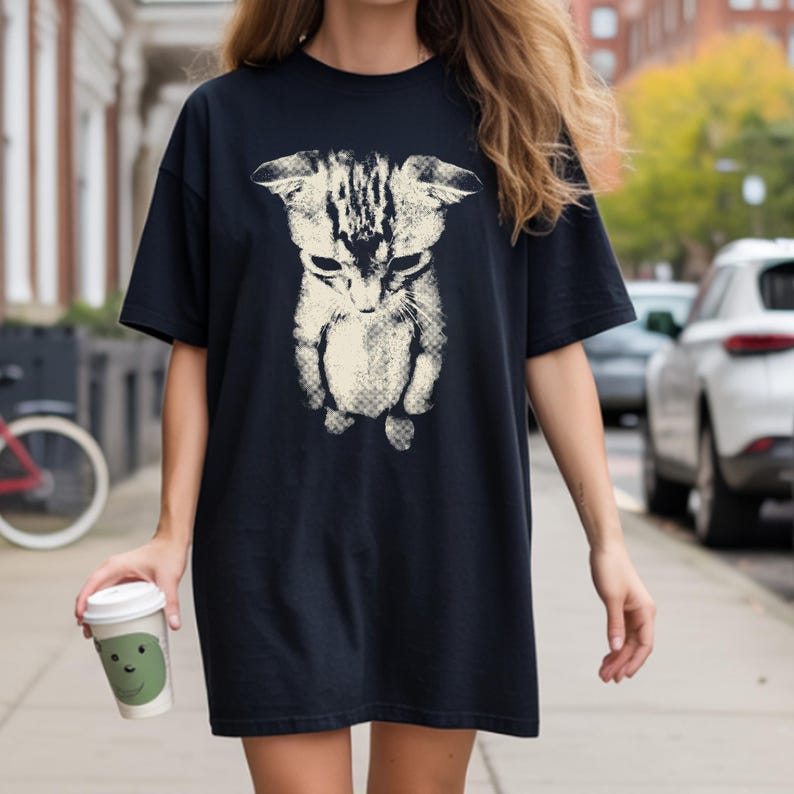 A high-contrast, vintage halftone graphic design on a dark shirt featuring the viral 'sad cat' meme image. The print depicts a small, cream-colored tabby kitten looking downward with a melancholy expression, flattened ears, and a sad face. The artwork has a distinct retro screentone texture with visible dot patterns, giving it a stylized, grunge-inspired look