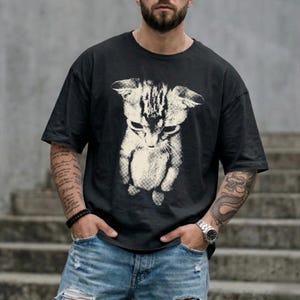 Peut inclure: Un t-shirt noir &agrave; manches courtes avec une grande impression graphique blanc cass&eacute; repr&eacute;sentant un chaton &agrave; l'aspect textur&eacute; et vieilli. Le chaton est repr&eacute;sent&eacute; en position debout, la t&ecirc;te inclin&eacute;e vers le bas, centr&eacute; sur le devant.