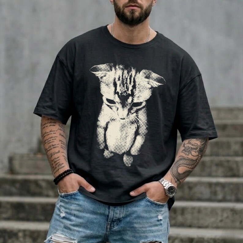 Peut inclure: Un t-shirt noir &agrave; manches courtes avec une grande impression graphique blanc cass&eacute; repr&eacute;sentant un chaton &agrave; l'aspect textur&eacute; et vieilli. Le chaton est repr&eacute;sent&eacute; en position debout, la t&ecirc;te inclin&eacute;e vers le bas, centr&eacute; sur le devant.
