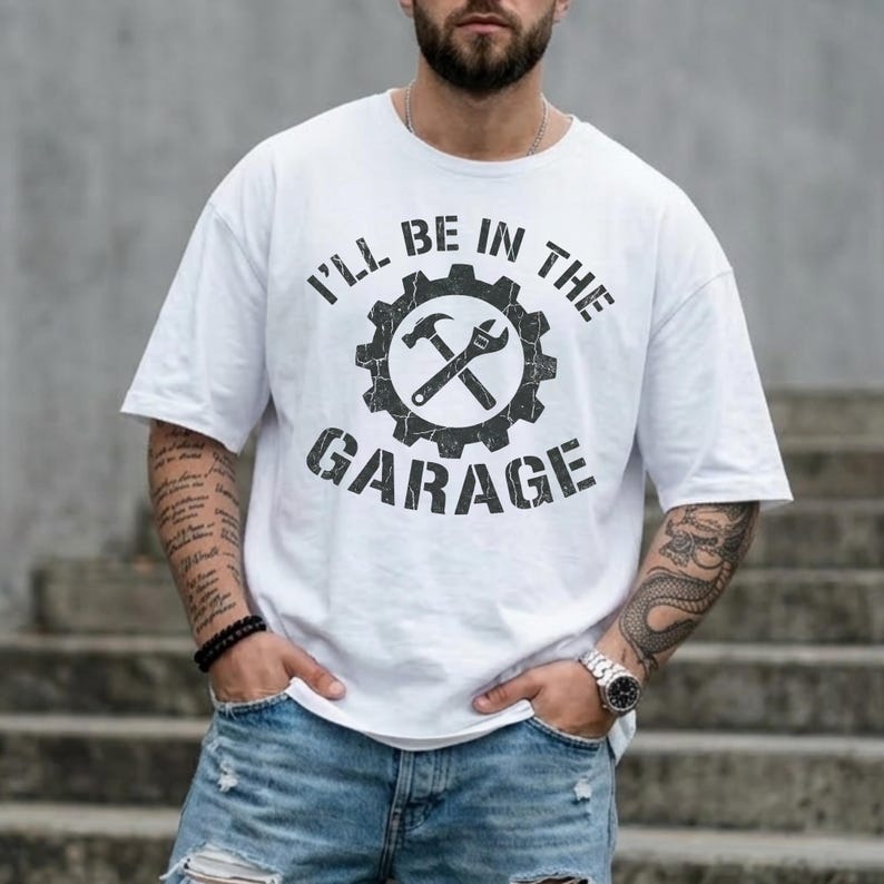 May include: White t-shirt with the text "I'll Be In The Garage" in a distressed black font. The design features a gear with a hammer and wrench crossed inside. The shirt is a casual style.