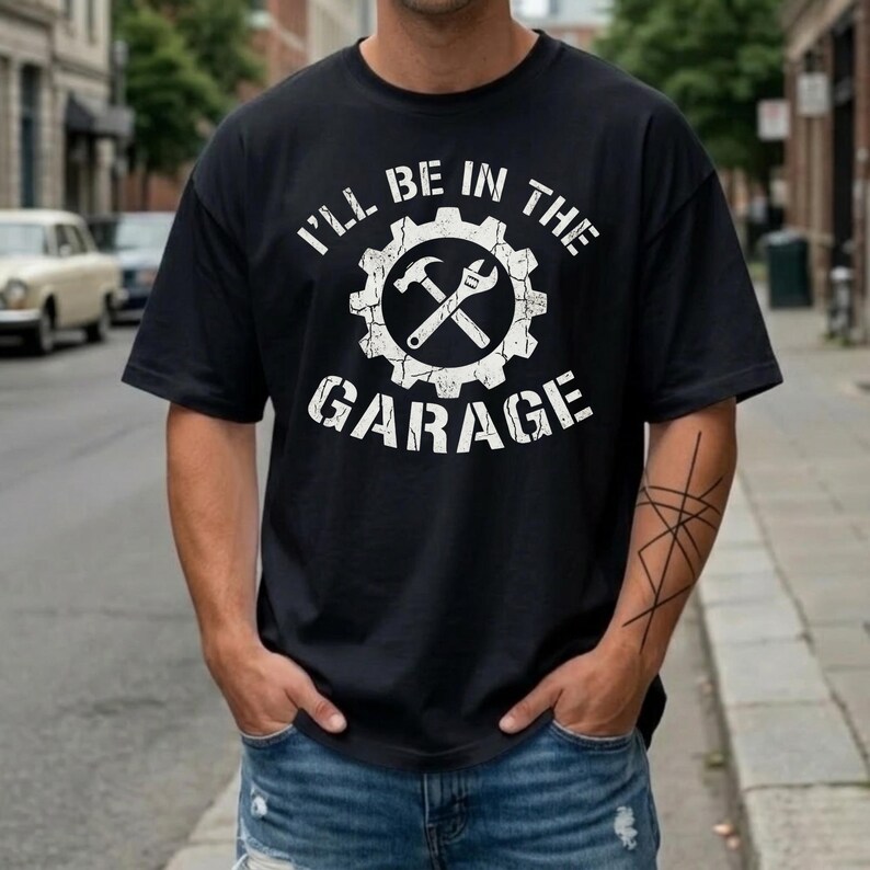 A rugged, industrial-style graphic featuring the text "I'LL BE IN THE GARAGE." The design centers on a large gear icon containing a crossed hammer and adjustable wrench. The entire white graphic has a heavily distressed, weathered texture, giving it a vintage workshop feel. The text is arched around the central emblem, and the design is centered on a black t-shirt for a high-contrast, masculine look.