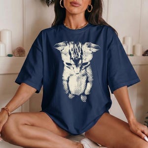 Peut inclure: Un t-shirt bleu marine &agrave; manches courtes avec une grande impression graphique couleur cr&egrave;me repr&eacute;sentant un chat sur le devant. L'illustration du chat pr&eacute;sente un aspect textur&eacute; et us&eacute; avec des motifs de hachures visibles.