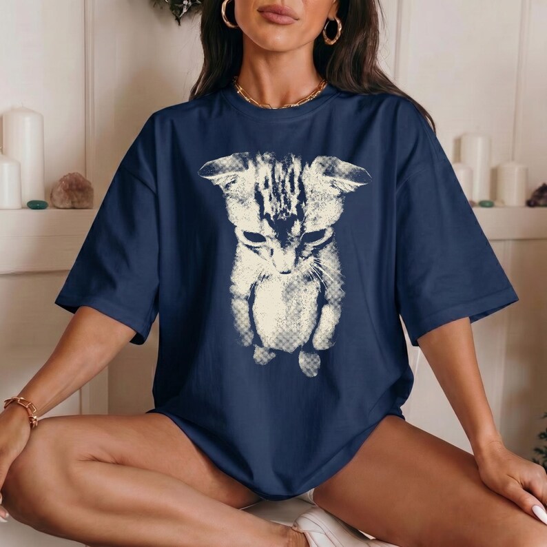 Peut inclure: Un t-shirt bleu marine &agrave; manches courtes avec une grande impression graphique couleur cr&egrave;me repr&eacute;sentant un chat sur le devant. L'illustration du chat pr&eacute;sente un aspect textur&eacute; et us&eacute; avec des motifs de hachures visibles.