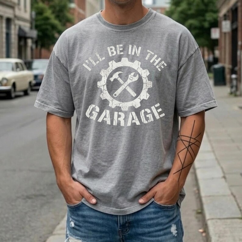 May include: Gray t-shirt with the text "I'LL BE IN THE GARAGE" in white. The design includes a gear, hammer, and wrench. The shirt is worn with blue jeans.