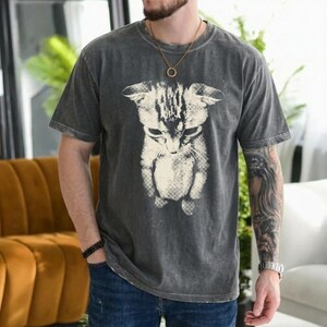 Peut inclure: Un t-shirt &agrave; manches courtes gris anthracite avec une grande impression graphique blanc cass&eacute; repr&eacute;sentant un chat regardant vers le bas. Le tissu pr&eacute;sente une texture d&eacute;lav&eacute;e de style vintage. Le t-shirt a un col rond et une coupe d&eacute;contract&eacute;e.