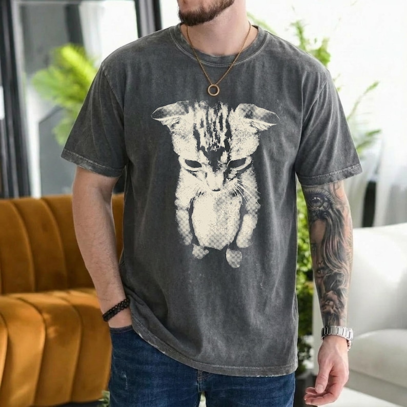Peut inclure: Un t-shirt &agrave; manches courtes gris anthracite avec une grande impression graphique blanc cass&eacute; repr&eacute;sentant un chat regardant vers le bas. Le tissu pr&eacute;sente une texture d&eacute;lav&eacute;e de style vintage. Le t-shirt a un col rond et une coupe d&eacute;contract&eacute;e.