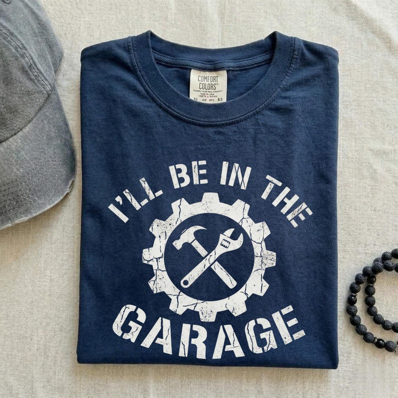 May include: Navy blue t-shirt with the text "I'll Be In The Garage" in white. The design includes a gear, hammer, and wrench. A gray baseball cap and a black beaded bracelet are also in the image.