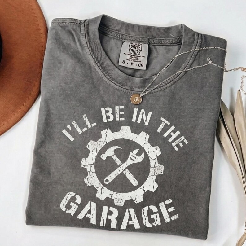 May include: Gray Comfort Colors t-shirt with the text "I'll Be In The Garage" in white. The design includes a gear, hammer, and wrench graphic. A silver necklace with a pendant is draped on the shirt.