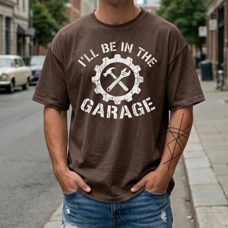 May include: Brown t-shirt with the text "I'll be in the garage" in white. The graphic features a gear with a hammer and wrench crossed inside. The shirt is worn with blue jeans.