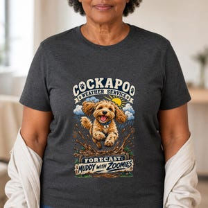 Puede incluir: Una camiseta gris marengo jaspeado con un gráfico de un perro Cockapoo de color marrón dorado saltando en el barro. El texto dice COCKAPOO WEATHER SERVICE y FORECAST: MUDDY WITH ZOOMIES, junto a ilustraciones de un sol, nubes y huellas.