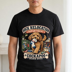 May include: A black t-shirt featuring a graphic of a brown Cockapoo dog holding a striped sock in its mouth. The text reads SOCK RELOCATION DEPARTMENT and COCKAPOO BRANCH OFFICE, with illustrations of a green filing cabinet and a basket of socks.