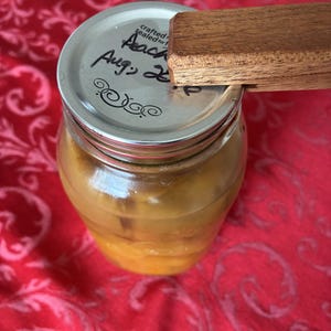 Może przedstawiać: Szklany słoik wypełniony żółtym przetworem, zamknięty metalową pokrywką i drewnianym narzędziem. Na pokrywce napis "Peach Aug 2". Słoik stoi na czerwonej tkaninie we wzory.