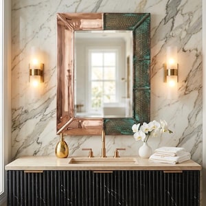 Puede incluir: Un espejo rectangular con marco de cobre y borde texturizado verde cuelga sobre un mueble de baño. El mueble tiene un armario negro acanalado, una encimera beige y accesorios dorados. Dos apliques flanquean el espejo. Un jarrón blanco con flores está en la encimera.