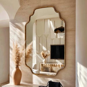 May include: A large, ornate mirror with a gold-colored frame hangs on a textured wall. The mirror has a unique, scalloped shape. A vase with dried pampas grass sits to the left of the mirror. A black purse rests on a surface below the mirror.